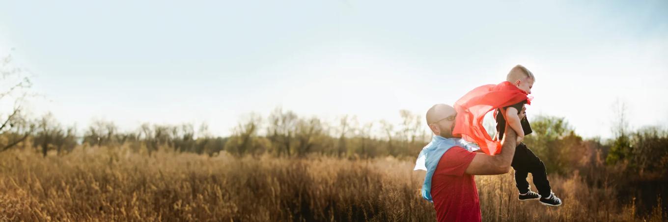Man with son playing outside wearing capes like super hero Superman. Happily playing outside in a field.