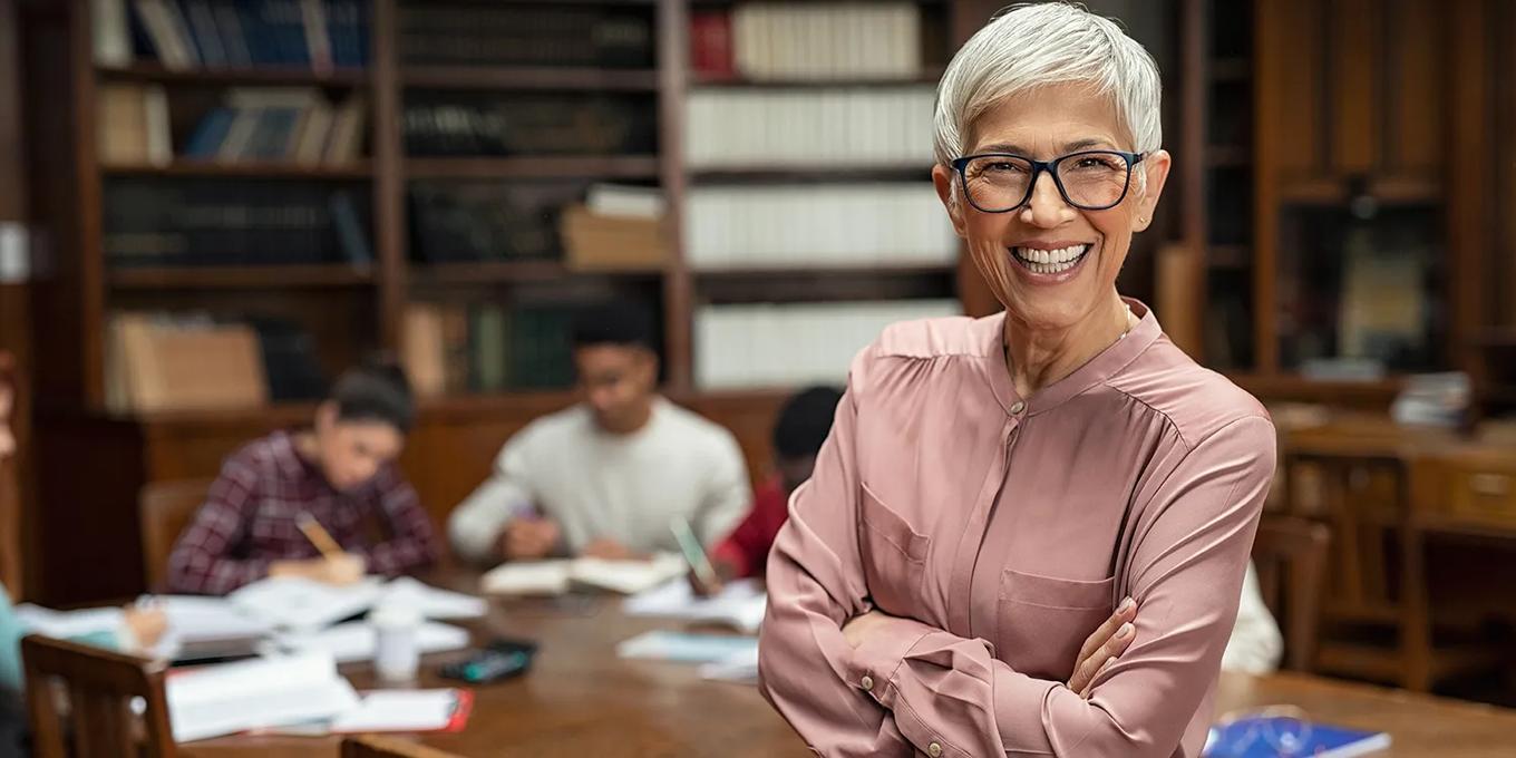 Portrait of mature professor with crossed arms standing in university library and looking at camera with copy space. Happy senior woman at the library working as a librarian. Satisfied college teacher smiling with students in background studying.