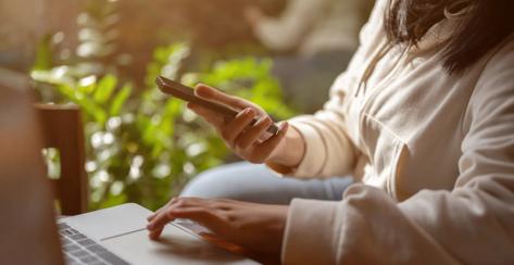 Cropped image of an Asian woman holding her smartphone while typing on her laptop keyboard, working at a table indoors. People and technology concepts