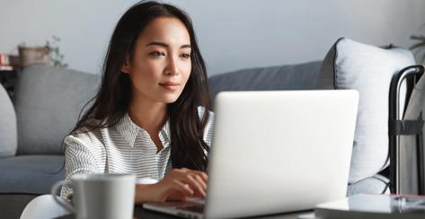 Young ambitious asian girl working remote from home, looking at laptop screen and smiling. Woman checking mail or researching while telecommuting, sitting on floor at her apartment.
