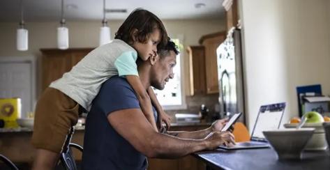 Father sitting at the kitchen table reviewing his benefits online on the computer with son looking over his shoulder,
