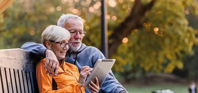 Smiling senior active couple sitting on bench looking at tablet computer.
