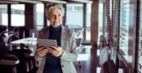Older lady standing at her desk looking out ithe window with an ipad in hand.,