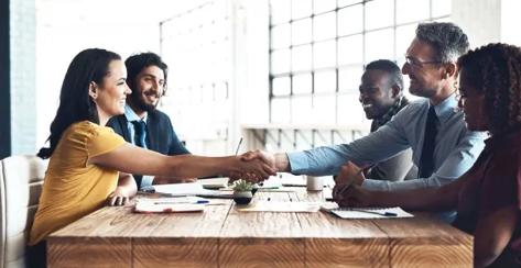 A group of five professionals meeting in a modern office as two of them shake hands across a wooden conference table.