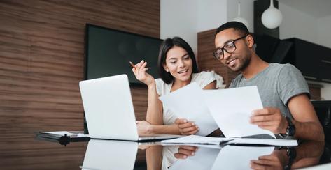 Photo of cheerful loving young couple using laptop and analyzing their finances with documents. Look at papers.