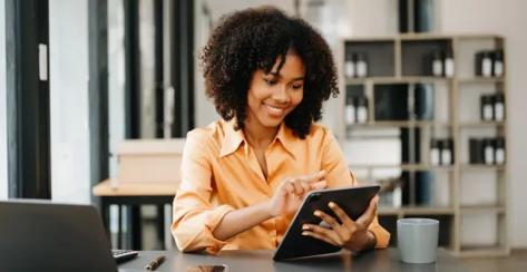 Black woman wearing an orange shirt smiles as she uses a tablet at her desk.