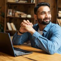 Happy and optimistic Indian man in casual wear using laptop in modern office, successful inspired arabic male entrepreneur looks away and smiles thoughtfully, planning next step
