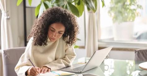 Woman writing in a journal sitting in front of her laptop.