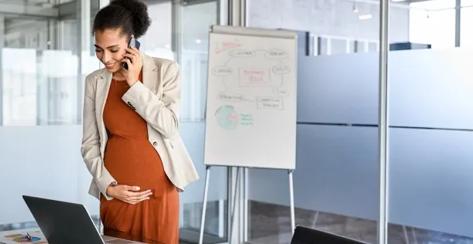 A Black, pregnant professional woman stands in a workplace conference room while talking on the phone.