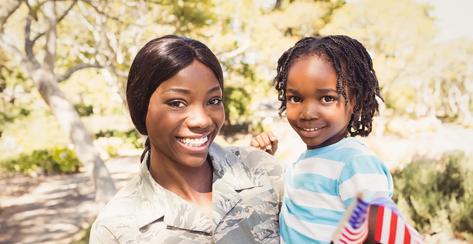 Young woman in a military uniform holding her young child who is smiling while waving an American flag