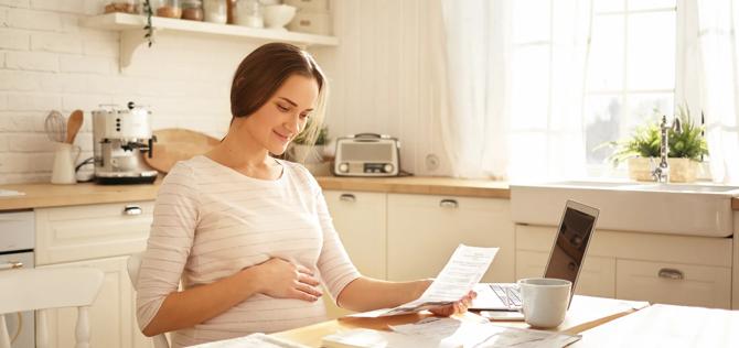 Beautiful young housewife expecting child calculating bills in kitchen, sitting at wooden table with laptop and papers