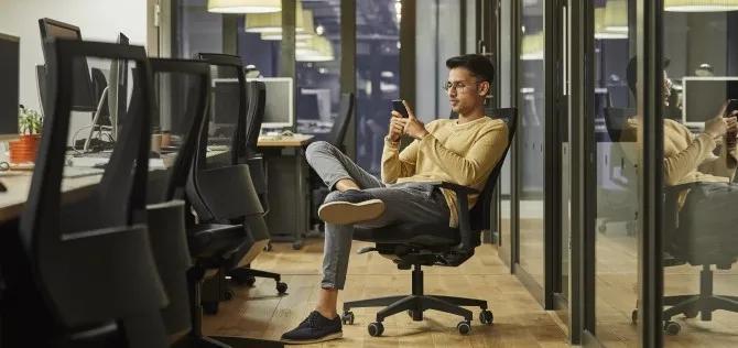 Young male employee sitting at his desk looking at his phone reviewing his benefits,