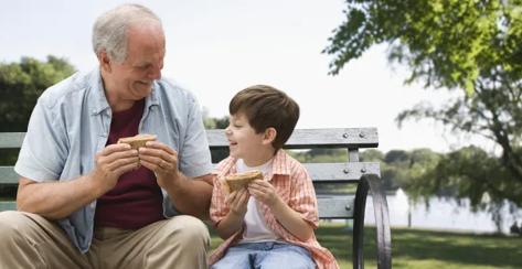 Grandfather and grandson sitting on a park bench eating sandwiches.