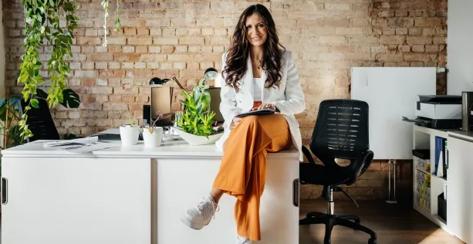 A confident woman in casual business attire with her legs crossed sits on a desk in a modern office