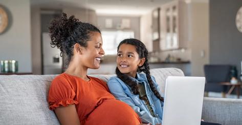 Pregnant black woman working on laptop while sitting on sofa and smiling to daughter. Cheerful expecting mother and girl relaxing on couch and using laptop to book vacation. Happy woman with baby bump and daughter working on computer from home during maternity leave.