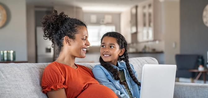 Pregnant black woman working on laptop while sitting on sofa and smiling to daughter. Cheerful expecting mother and girl relaxing on couch and using laptop to book vacation. Happy woman with baby bump and daughter working on computer from home during maternity leave.