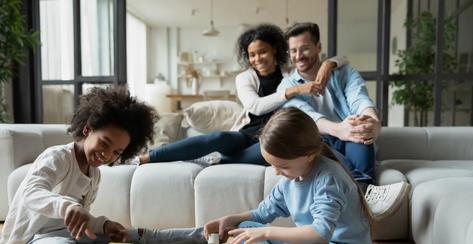 Happy bonding loving mixed race couple parents relaxing on comfortable sofa, watching adorable african american daughter playing toys with european sister on floor carpet in modern living room.