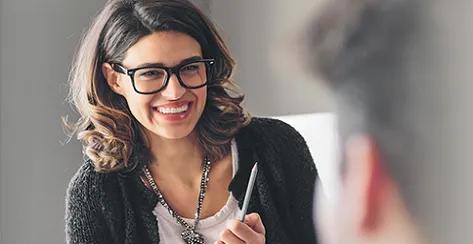 Woman with glasses and pen smiling at colleague.