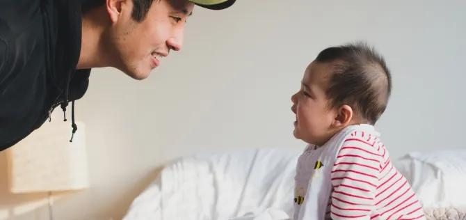 Father facing and engaging baby who is sitting on bed 