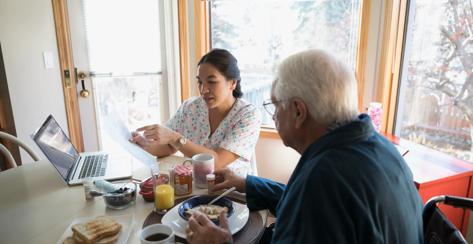 Home caregiver explaining paperwork to senior man eating breakfast at dining table