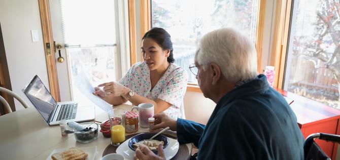 Home caregiver explaining paperwork to senior man eating breakfast at dining table