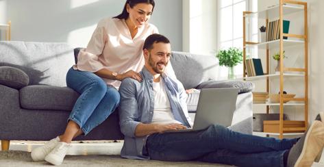 Young married couple sitting on sofa in the living room at home using tablet PC for internet and social media. Happy family resting on couch enjoying weekend watching video or talking on video call.