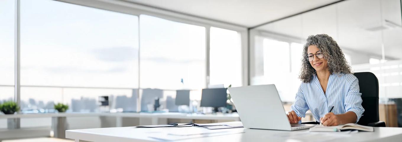 Busy mature business woman working in office using laptop writing. Smiling middle aged professional executive looking at computer, elearning, watching webinar having hybrid meeting sitting at desk.