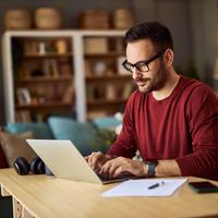 A focused male freelance content creator with glasses sitting at a desk and typing on his laptop while working from home.