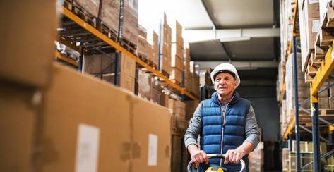 Senior male warehouse worker pulling a pallet truck with boxes.